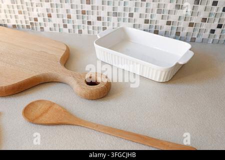 White ceramic baking dish with wooden cutting board and spoon on kitchen counter. Stock Photo