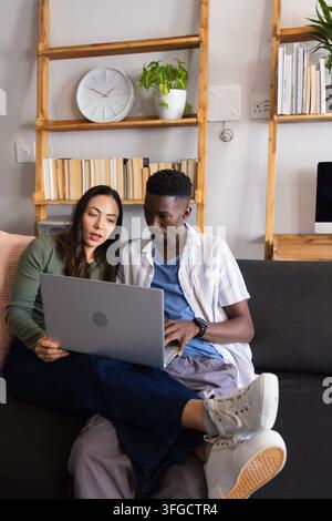 Couple sitting on dark gray couch using silver laptop, female wearing green top and white sneakers Stock Photo