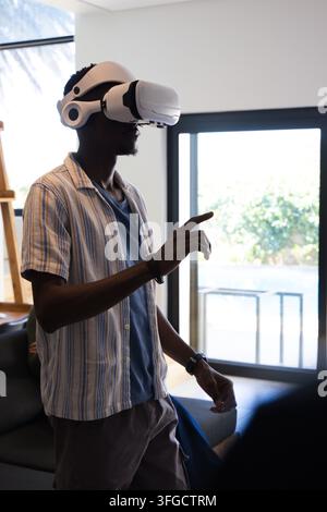 African American man wearing VR headset and striped shirt, pointing and interacting in living room Stock Photo