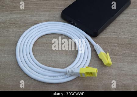 Close-up shot of a neatly coiled white network cable with yellow connectors paired next to a modern external storage device on a clean wooden table, s Stock Photo
