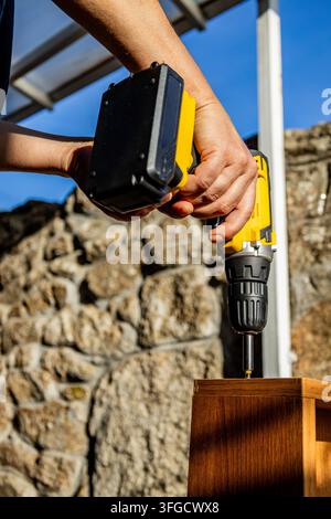 Close-up of a person using a yellow cordless drill to assemble a wooden cabinet outdoors beside a stone wall on a sunny day. Stock Photo