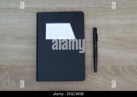 Flat lay view of a black notebook with a blank white label on the cover, accompanied by a sleek black pen on a light wood background. Stock Photo