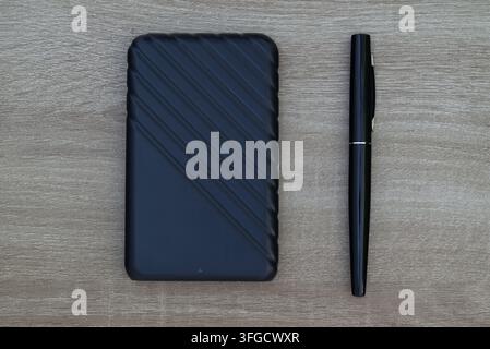 op-down view of a textured black external hard drive paired with a sleek black pen on a light wooden background. Stock Photo