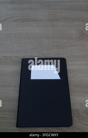 Vertical composition of a closed black notebook positioned at the bottom with ample negative space on a light wooden background. Stock Photo