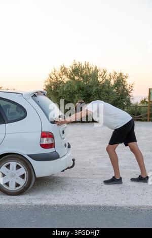 Young man putting effort pushing his broken down car on a dirt road, experiencing vehicle trouble and frustration Stock Photo
