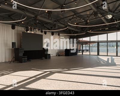 Bright daytime view inside modern event hall with circular ring lights overhead, stage equipment, tall white curtains, and wide glass walls showing ci Stock Photo