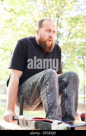 Bearded caucasian man doing dips on metal bars in sunlit park, face showing effort and concentration, wearing black tshirt and patterned jeans, urban fitness scene emphasizing strength and raw energy. Stock Photo