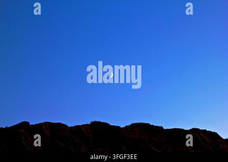 Crescent moon over silhouetted rocky hills at twilight against deep blue sky. Stock Photo