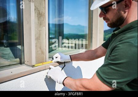 Construction worker measuring window frame with tape, checking dimensions before installation. Exterior finishing, accuracy control, renovation planning, professional building inspection concept Stock Photo