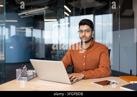 Young man with glasses works on laptop at desk inside modern glass office, wearing orange shirt beside tablet, notebook, pen holder, and notepad, in bright corporate workspace with professional Stock Photo