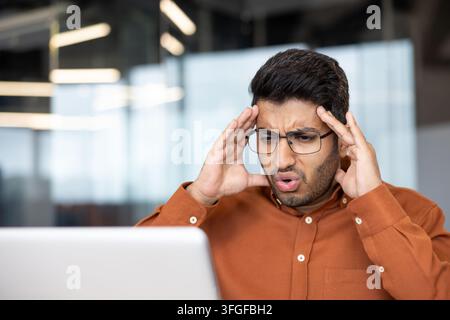 young man wearing glasses and rust shirt sits at desk with laptop, pressing temples with concerned expression, showing stress, focus, problem solving, and work pressure in modern office Stock Photo