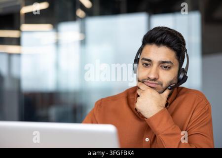 Focused young man in headset sits at laptop in bright office, resting chin on hand while concentrating on work, customer support, video call, or online collaboration in professional setting Stock Photo
