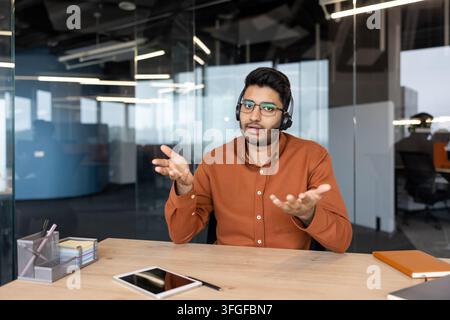 Young man wearing headset speaks and gestures during business video call in modern glass office meeting room, seated at desk with laptop, tablet, notebook, and stationery, conveying remote support Stock Photo