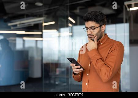 Thoughtful young man in glasses checks smartphone beside glass office wall, wearing orange shirt in modern workspace, with man, smartphone, office, business detail, showing thinking, focused, glass Stock Photo