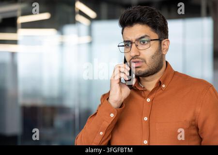 Worried office professional with orange shirt and glasses holds smartphone near glass wall, speaking with focused, serious expression in modern workspace, with man, glass, smartphone, mobile detail Stock Photo