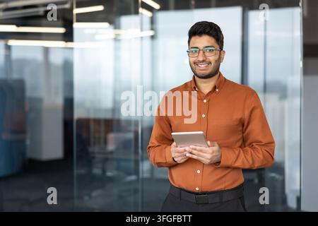 Portrait of young professional smiling while holding tablet in bright contemporary office hallway, with glass walls, soft reflections, and overhead lights, with man, smiling, glass, tablet detail Stock Photo