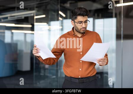 Young businessman in orange shirt studies printed documents inside modern office with glass partitions, holding separate pages while checking details under soft indoor light and calm professional Stock Photo
