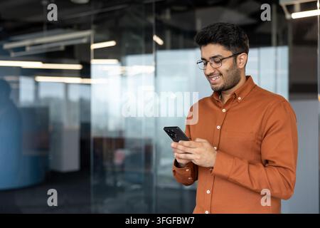 Smiling young man with glasses holds smartphone while standing in modern office interior with glass walls, natural light, and reflections, suggesting mobile messaging and connected workplace Stock Photo