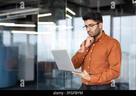 Young professional in orange shirt and glasses stands inside modern glass office, holding open laptop and resting hand on chin while reviewing work with focused concentration and calm posture Stock Photo