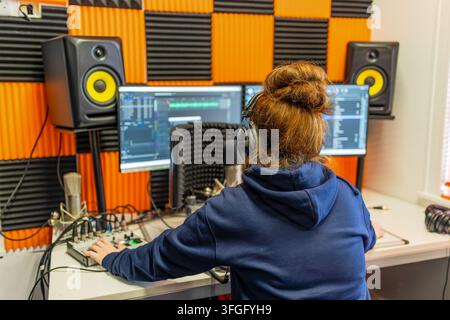 Red-haired woman wearing headphones operates audio mixer and recording software on dual monitors, surrounded by orange and black acoustic foam panels Stock Photo