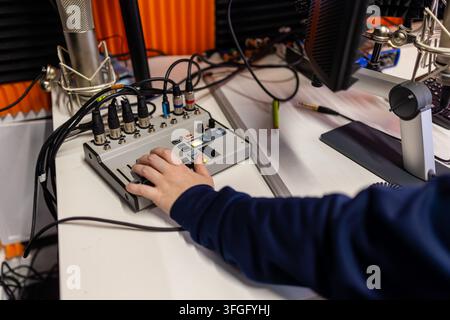 Close-up of hand in navy sleeve working faders and buttons on small broadcast mixer with multiple XLR cable inputs, condenser microphone and monitor v Stock Photo