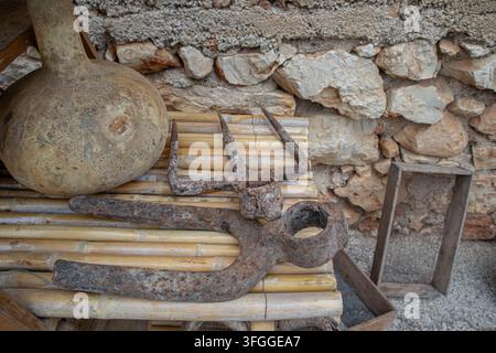 Rusty pitchfork heads and hand tools lie beside a dried bottle gourd calabaza on a wooden table near a stone wall. This scene shows vintage Spanish eq Stock Photo