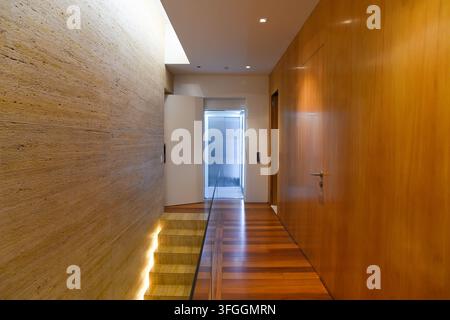 Modern Hallway Design: A contemporary hallway featuring warm wooden paneling, a unique textured wall, and illuminated staircase leading to an elevator Stock Photo