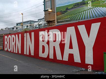 Bold red “Born in Bray” mural beside the DART railway in Bray, County Wicklow, Ireland, with a solar-energy billboard overlooking the line. Stock Photo