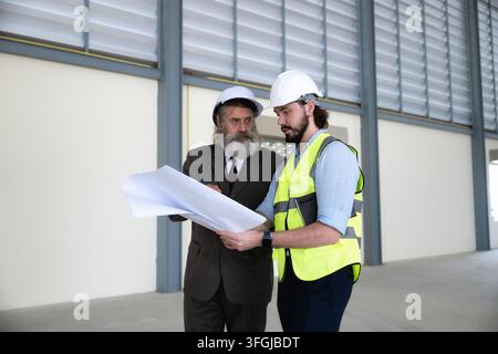 Engineer and senior businessman analyzing construction blueprint inside empty warehouse and reviewing architectural layout industrial building. Concep Stock Photo