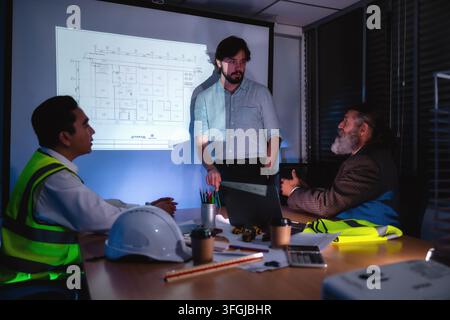 Engineering presenting and discuss floor plans on projector screen during late-night company meeting with senior project manager. Concept of brainstor Stock Photo