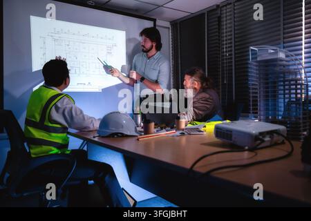 Engineering presenting floor plans on projector screen during late-night company meeting with senior project manager. Concept of brainstorming in cons Stock Photo