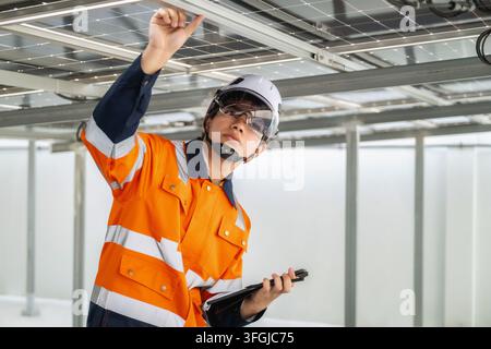 Young engineer in orange uniform and white helmet examines solar panels and metal beam structure reviewing installation details on tablet. expression Stock Photo