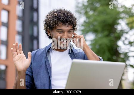 Friendly college student waving hand during online lecture webinar on laptop outdoors near university. Stock Photo