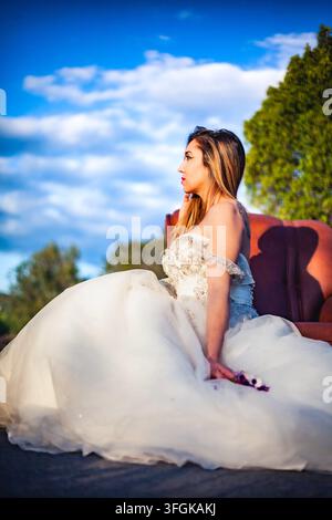 Bride in a white gown holding a bouquet while seated on a vintage red velvet sofa outdoors, Algarve, Portugal. Stock Photo
