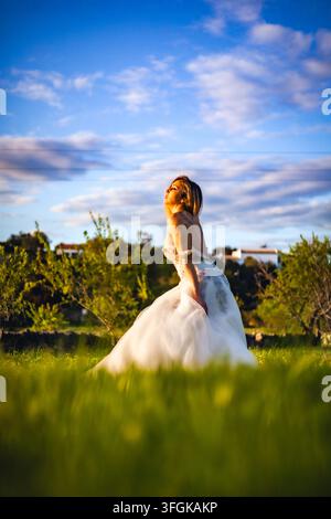 A bride in an elegant white wedding dress standing in a green meadow under a dramatic blue sky with clouds at sunset, in a rural countryside setting. Stock Photo