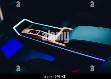 Modern car interior closeup of a sleek center console with ambient blue lighting, leather armrest, and electronic controls. Stock Photo