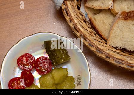 A top view shot of sliced white bread in a wicker basket next to pickled cucumbers and cherry tomatoes on a plate Stock Photo