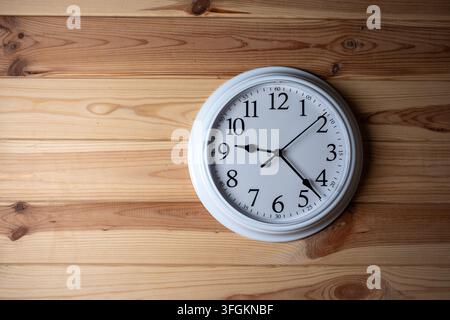 A round white analog wall clock with black arrows hanging on a light textured wooden background with copy space Stock Photo