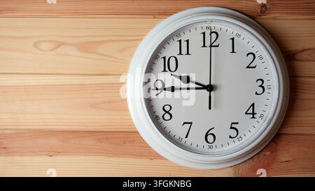 A classic white analog wall clock with black numbers hanging on a light textured wooden background with copy space Stock Photo