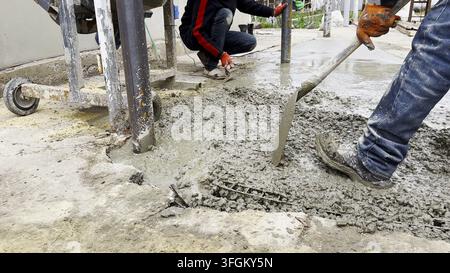 Male hands in protective gloves uses trowel to spread and level fresh cement in yard. Another worker distributes and moves the mixture with a shovel Stock Photo