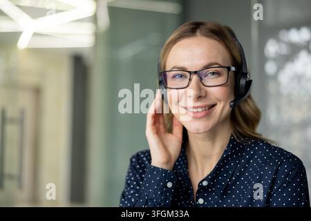 Customer support and remote communication, smiling female operator wearing headset and glasses working in modern office environment to provide professional assistance and help desk service solutions. Stock Photo