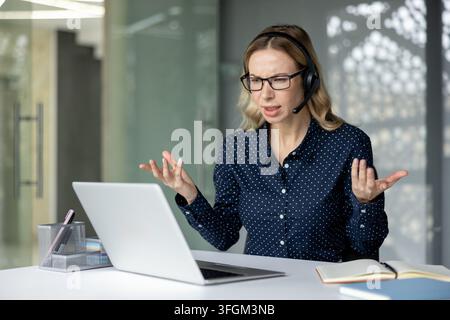 Customer support frustration and workplace stress, woman wearing headset working on laptop in office environment. Stock Photo