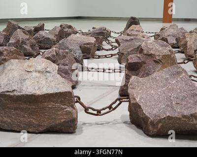 A close-up view of a contemporary art installation consisting of numerous large, rough-hewn rocks or boulders connected by heavy, rusted metal Stock Photo