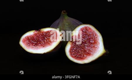 Two fresh figs are cut in half and placed on a dark surface. The inside shows a rich pink color with small seeds. The outside skin is dark purple with Stock Photo