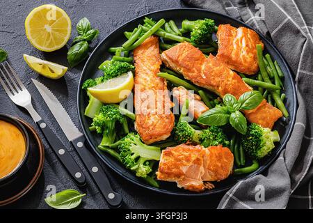 Grilled salmon portions with steamed green beans, broccoli florets, lemon, fresh basil on black plate over dark concrete table with cutlery and sauce, Stock Photo