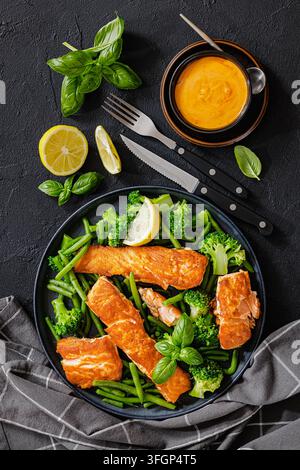 Grilled salmon portions with steamed green beans, broccoli florets, lemon, fresh basil on black plate over dark concrete table with cutlery and sauce, Stock Photo