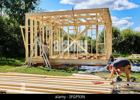 Construction crew assembles wooden frame with circular saw and lumber while one worker cuts boards and second secures bracing inside structure. Stock Photo
