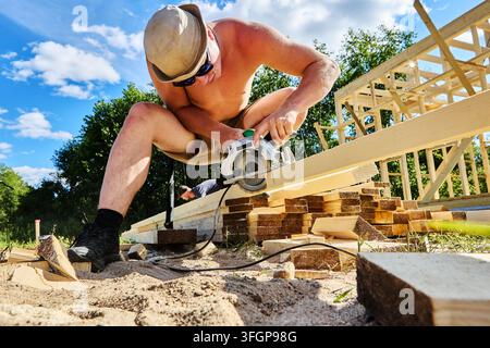 Operating circular saw on wooden board, young builder performs carpentry work during construction of house frame. Stock Photo