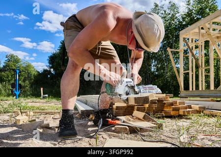 Using corded handheld circular saw with transverse motor to cut timber boards, young builder constructs wood frame house on construction site. Stock Photo