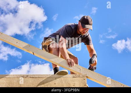 Securing timber roof framing using battery drill, young builder installs rafter on wooden construction frame. Stock Photo